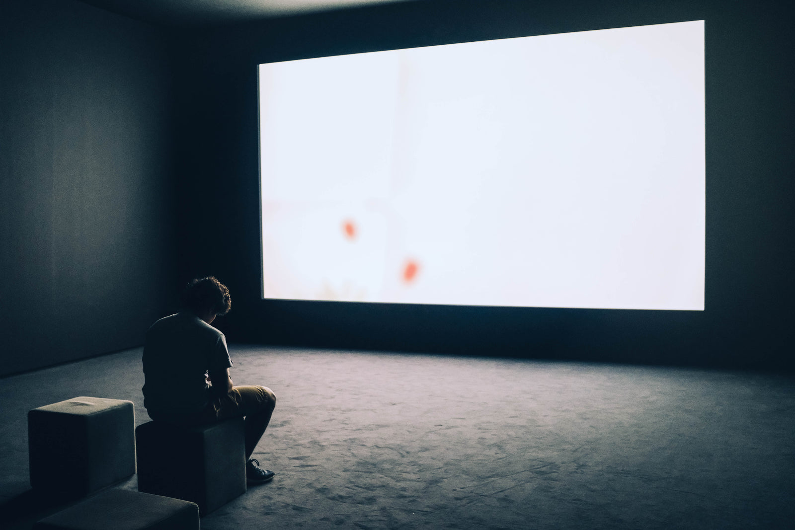 A lone viewer in a dark room facing a large, softly lit projection screen