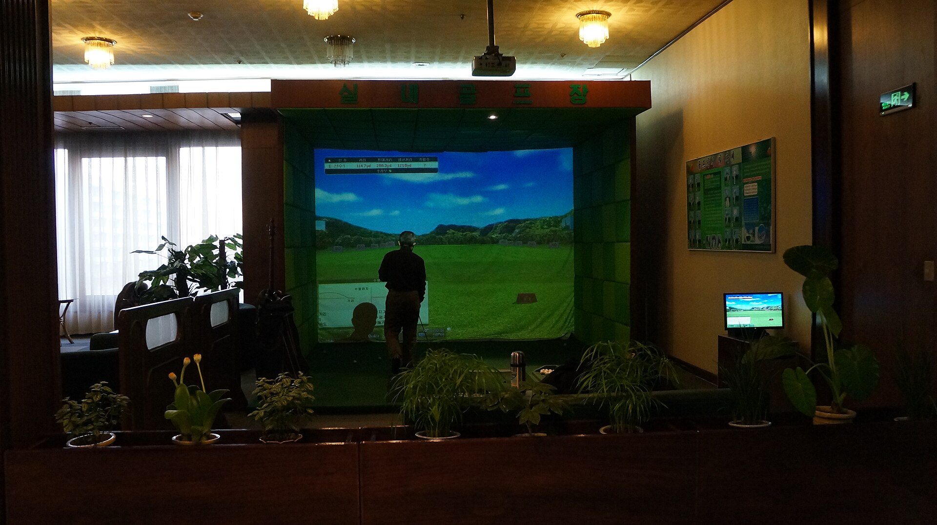 An indoor golf simulator bay in a plant-lined lounge: a ceiling projector throws a driving-range course onto a large impact screen with a shot-distance overlay, while a golfer takes a swing on the hitting turf and a club bag waits to the side.