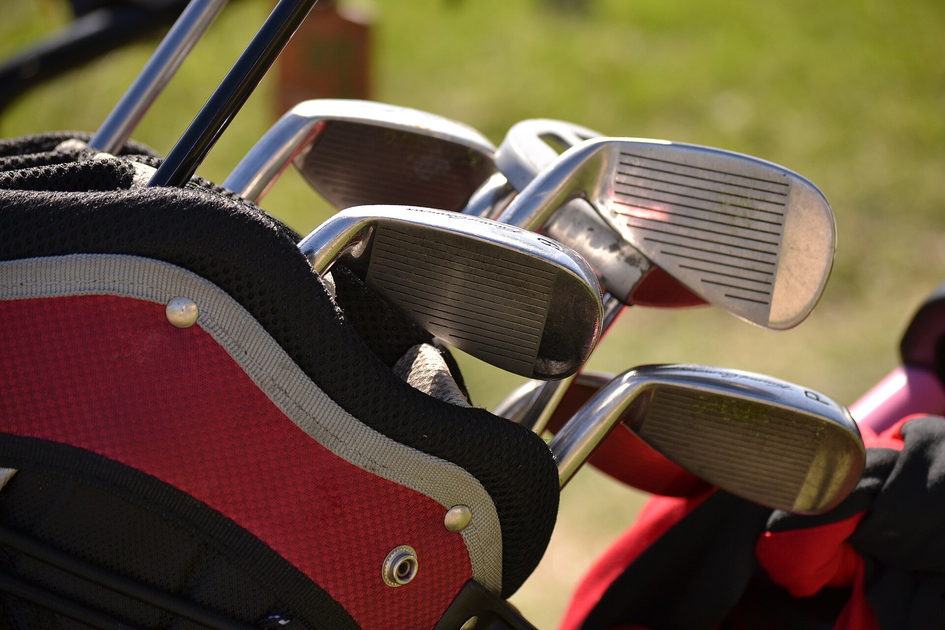 A close-up of a set of steel irons standing in a red-and-black golf bag on a grassy background.