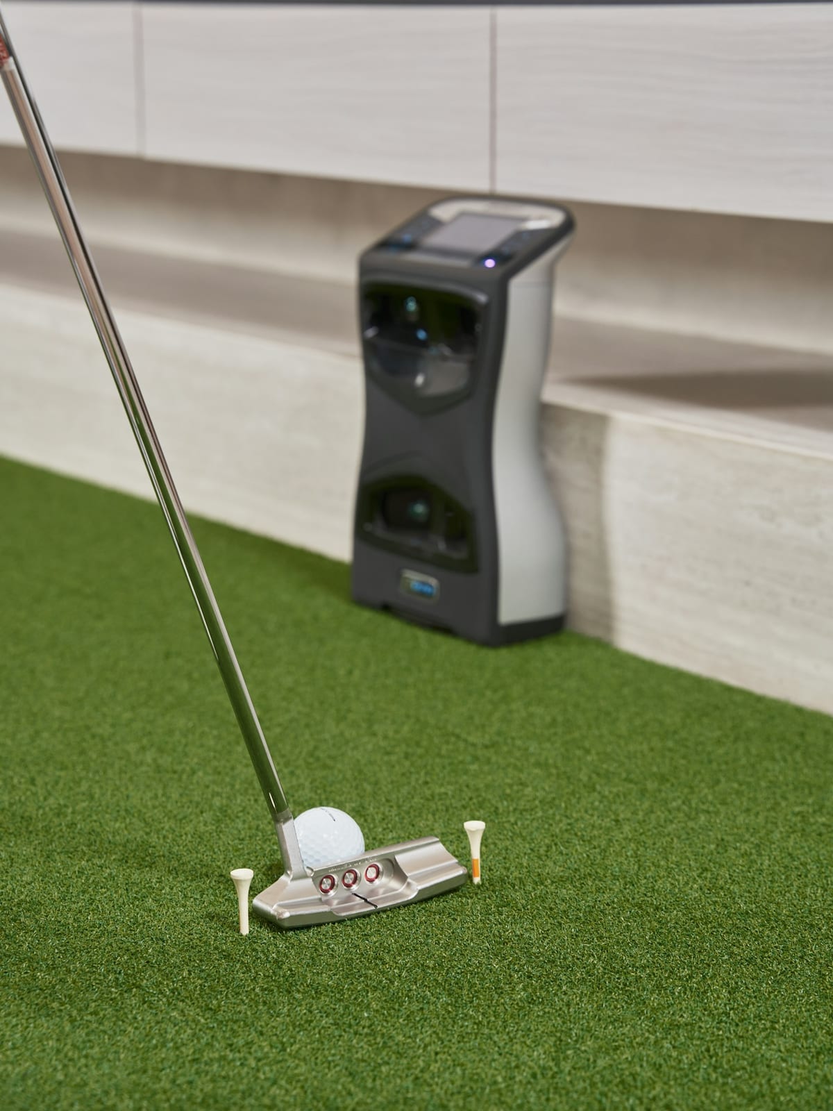 A dual-camera launch monitor on green hitting turf, a putter and ball in front of it.