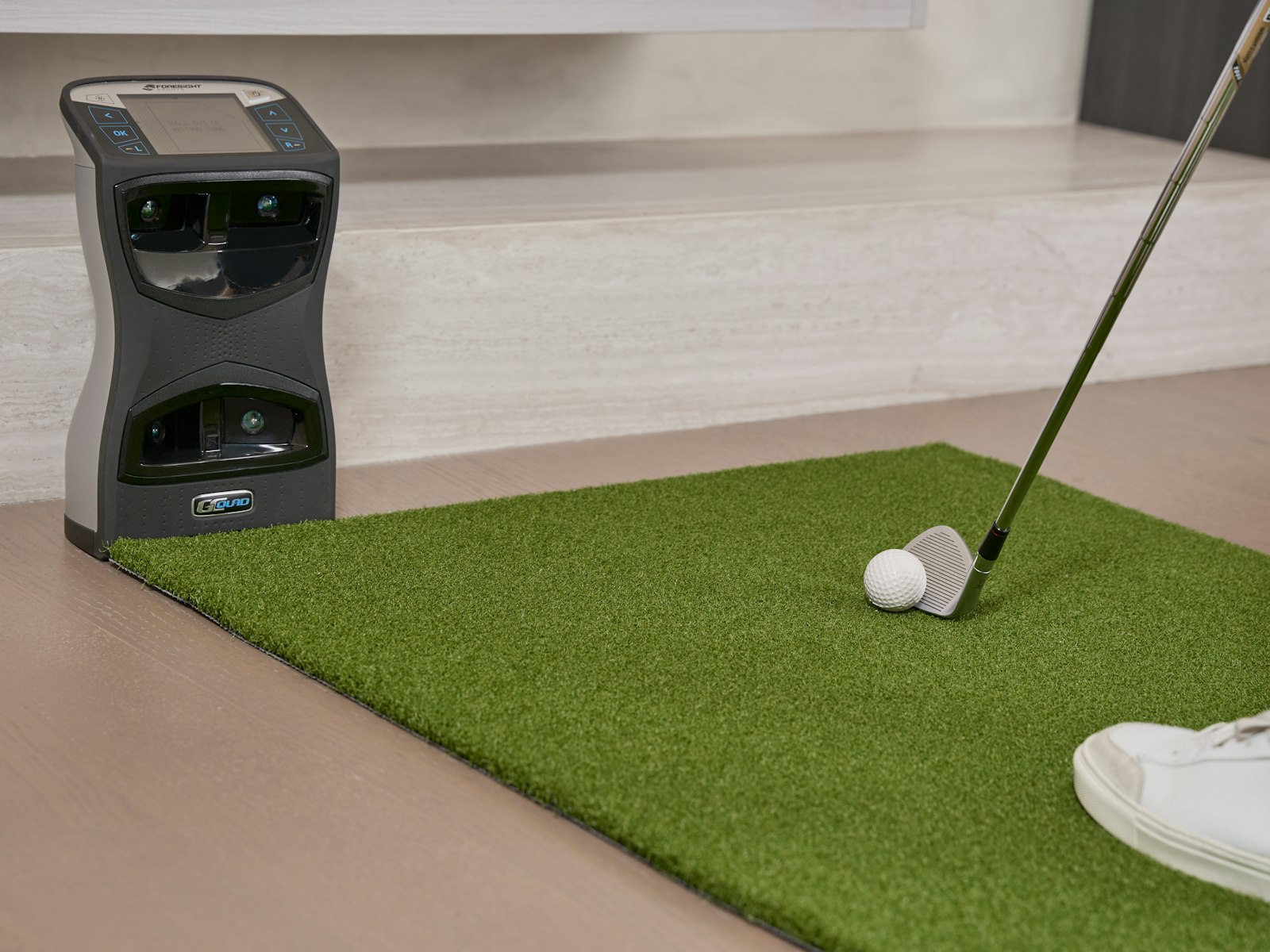 A launch monitor positioned beside a home chipping mat, capturing swing data indoors