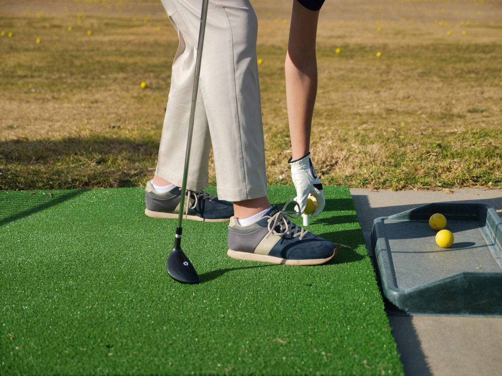 A golfer on a driving-range mat placing a yellow range ball on a tee with a gloved hand, a driver resting on the turf beside a tray of range balls.