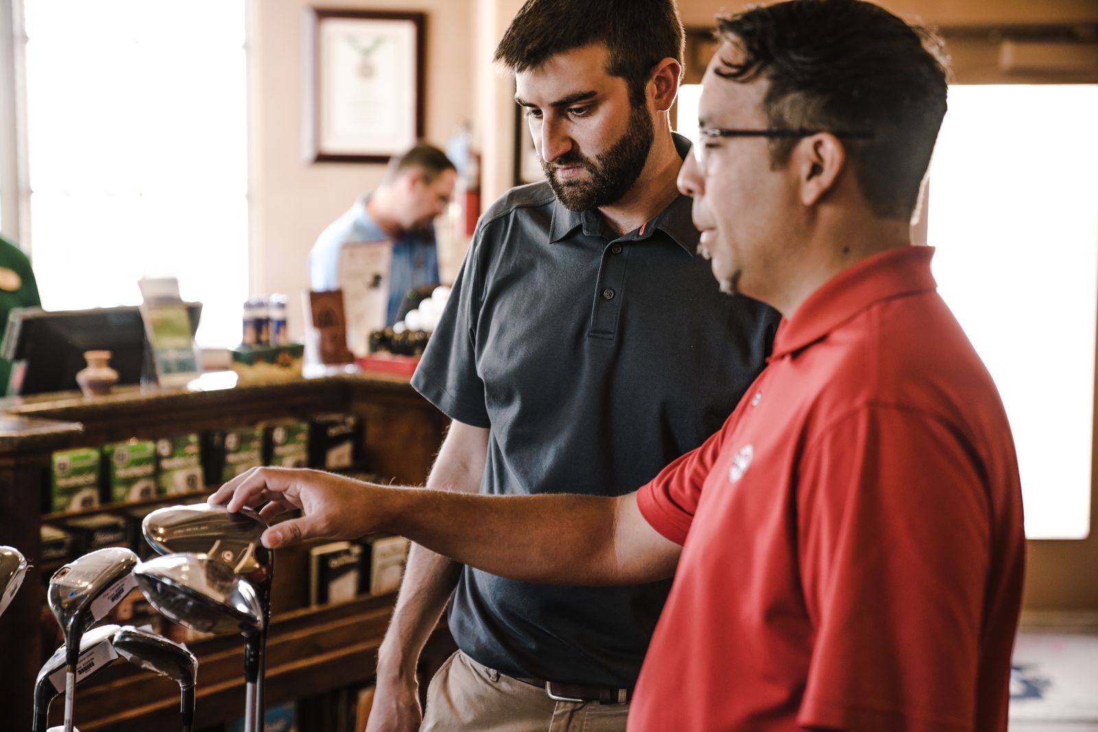 Two men in a golf pro shop lean over a rack of drivers and woods, one resting a hand on a clubhead, price tags hanging from the shafts.