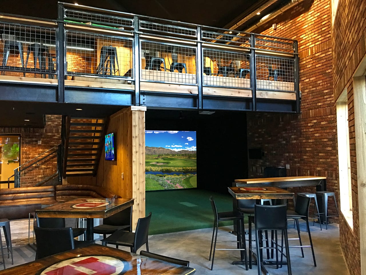 An unoccupied indoor golf simulator bay in a brick-walled lounge: a course hole projected on the impact screen, green hitting turf in front of it, and empty high-top tables to one side.