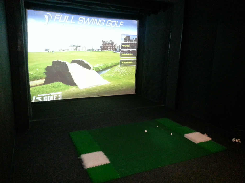 A home golf simulator bay with the hitting mat in the foreground and the impact screen several feet back, the mat carrying both left and right hitting strips