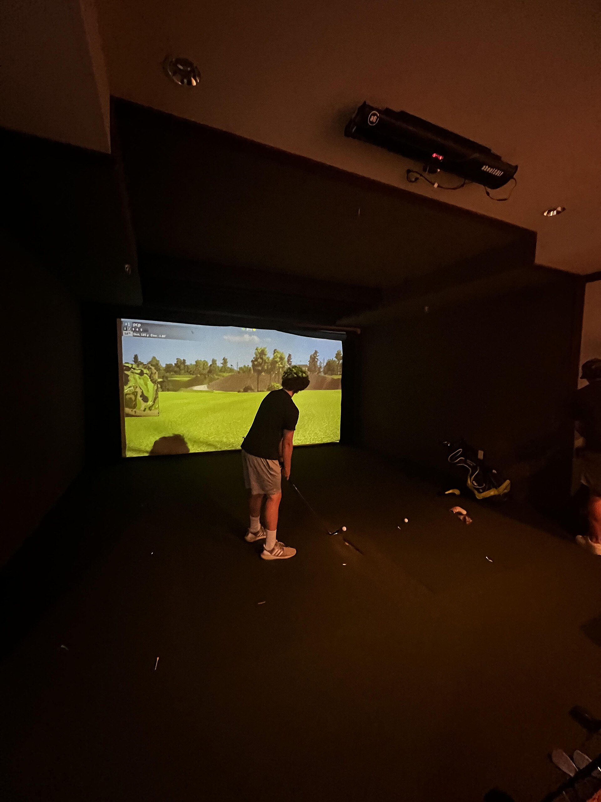 A golfer at address inside a home simulator bay with the projector mounted on the ceiling directly overhead